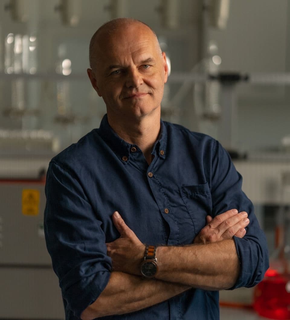 Man in a blue shirt with arms crossed, standing in a laboratory setting with blurred equipment in the background.